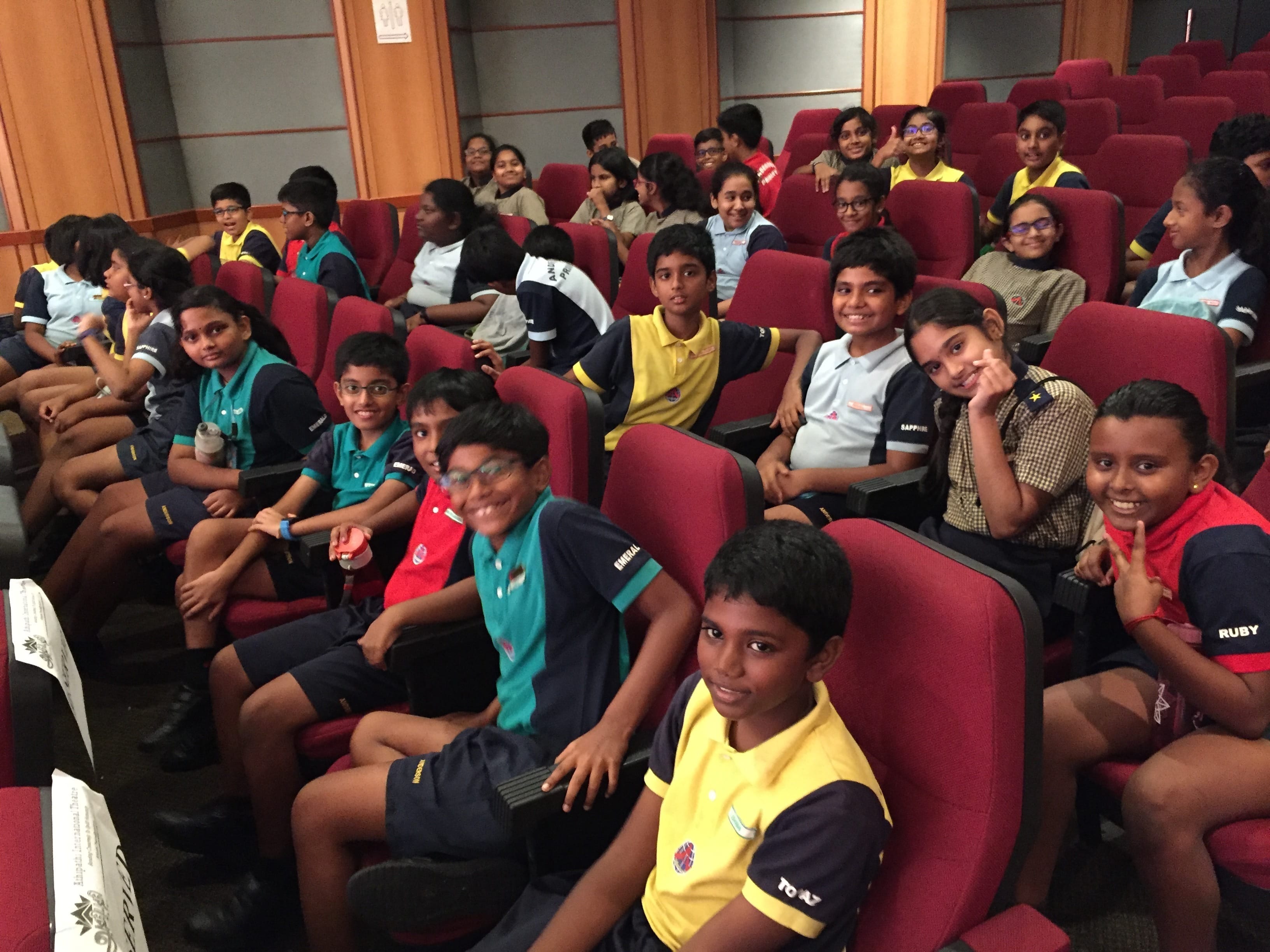 A large group of school children in various uniforms sitting in red auditorium seats, smiling at the camera.